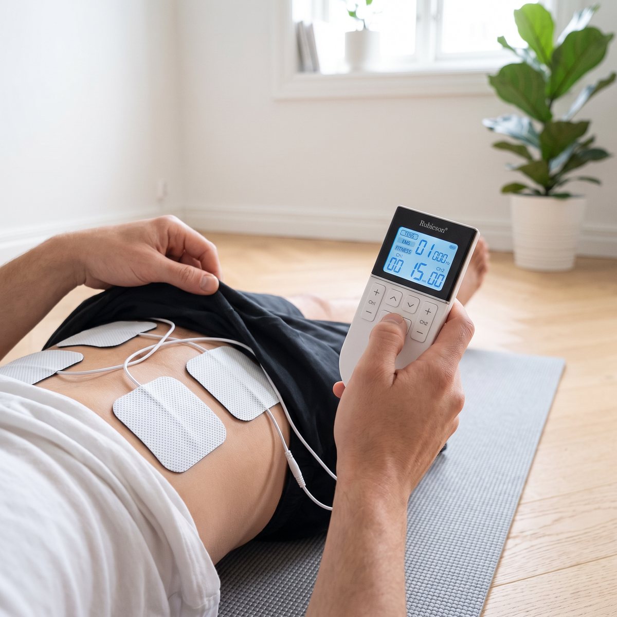 Rubicson TENS/EMS-dosa i handen med fyra elektrodplattor på magen, liggande på en yogamatta