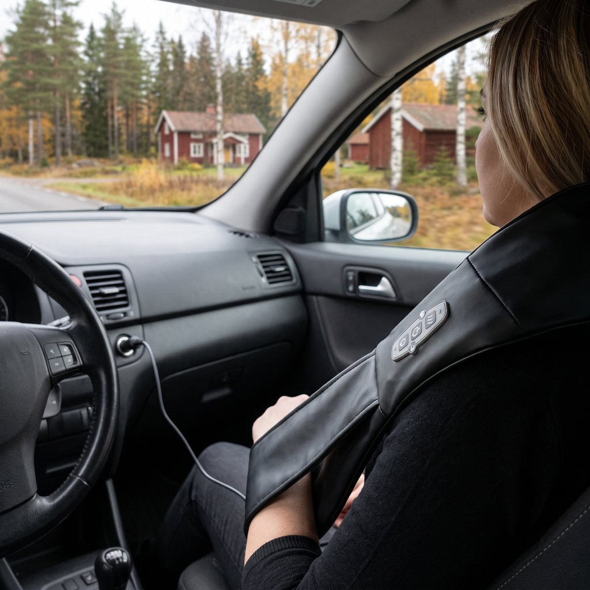 Svart Extrasim HomeTherapist över axlarna på en passagerare i bil, kopplad till 12-voltsuttaget, med röda stugor och höstskog utanför