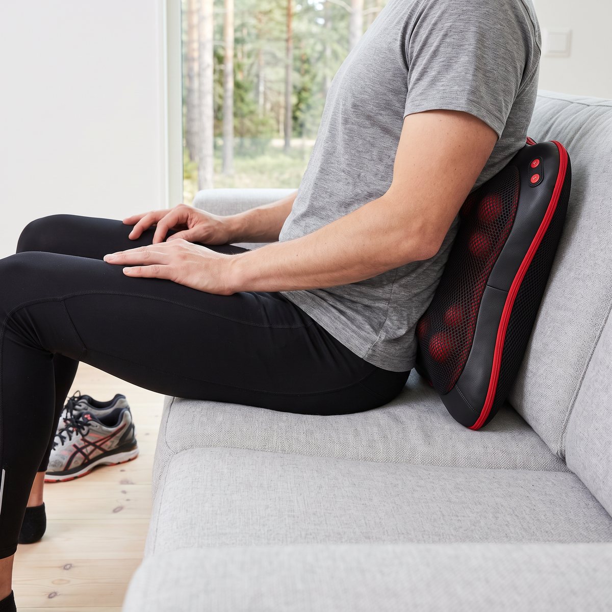 Svart Flowpillow Heat med röd kant bakom ländryggen på en person i löpartights i en ljus svensk soffa, med tallskog utanför