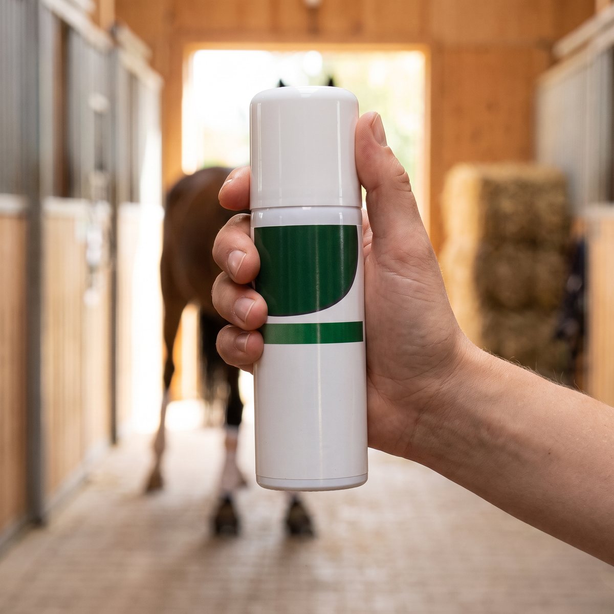 Vit sprayburk hålls i handen i en svensk stallgång med en häst i bakgrunden