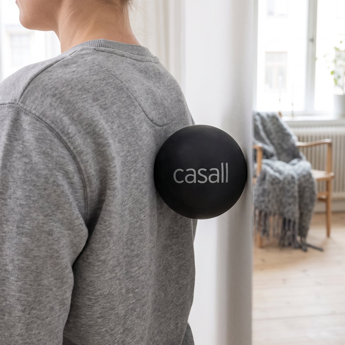 Svart massageboll av gummi från Casall mellan övre ryggen och en vit vägg i ett ljust svenskt vardagsrum