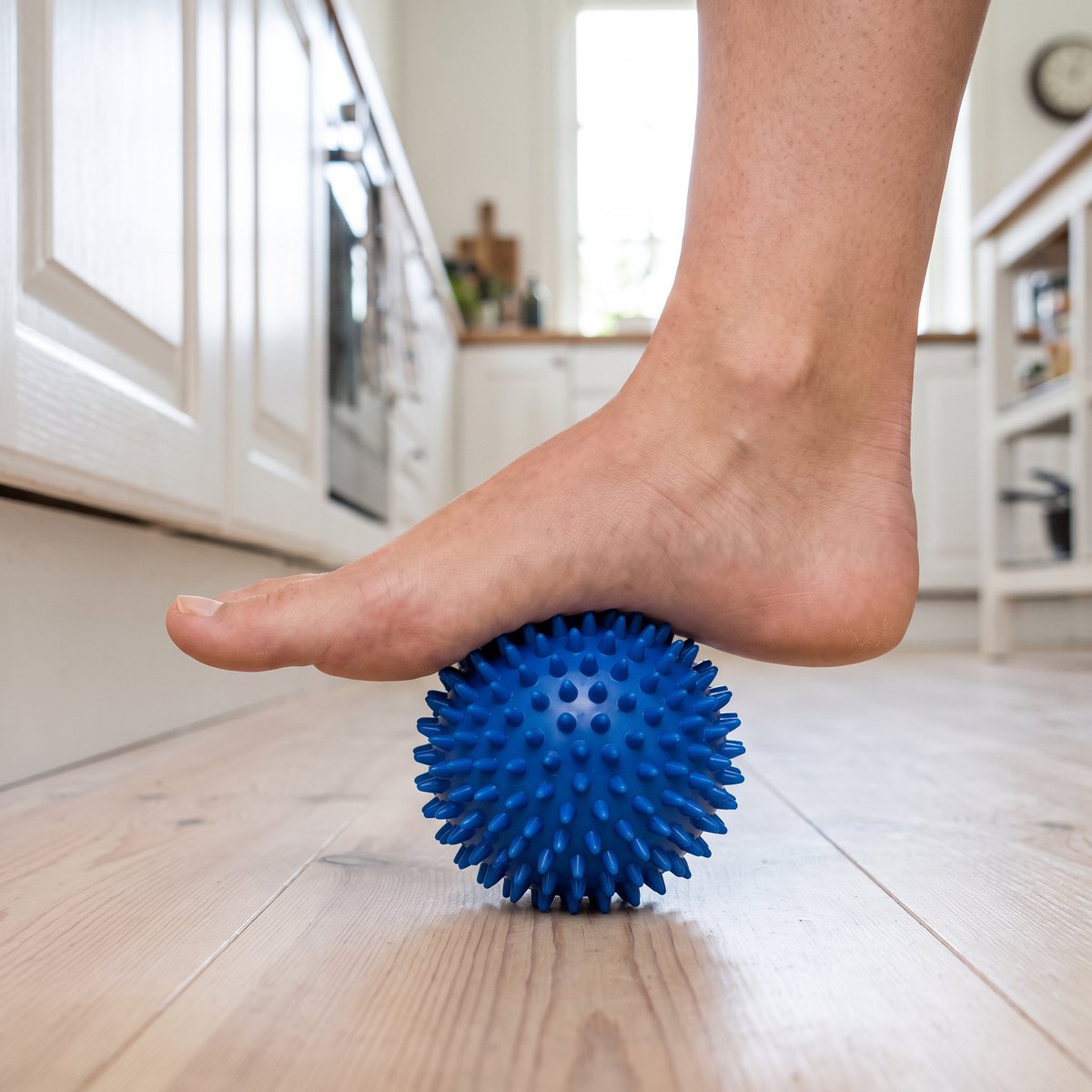 Blå massageboll med små piggar under foten hos en person som står vid en vit köksbänk i ett svenskt hem