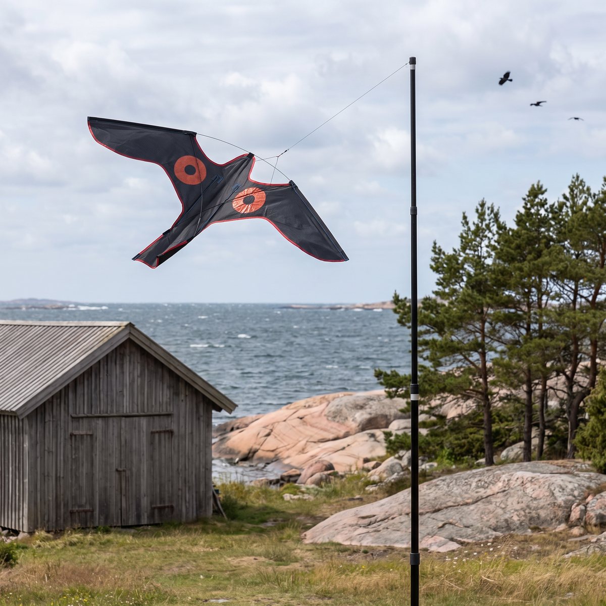 Svart rovfågelsdrake med röda ögonringar som flyger i en lina från en svart mast vid ett båthus och granithällar i skärgården