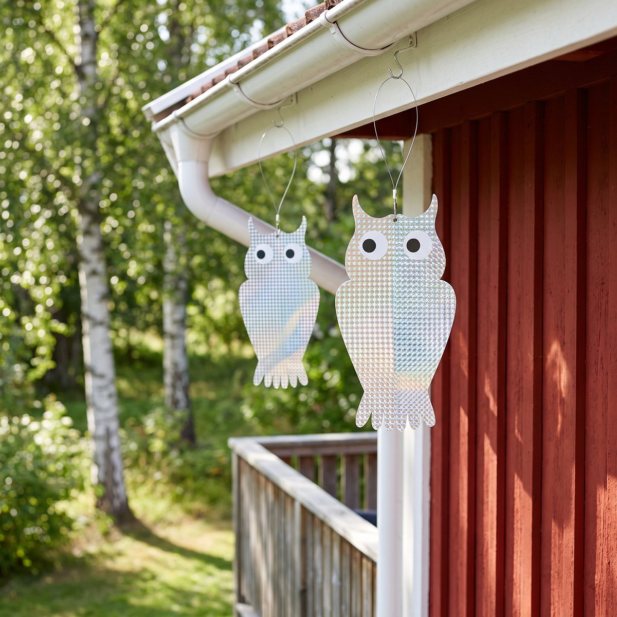 Två platta reflexugglor som hänger i krokar under stuprännan på ett rött svenskt trähus