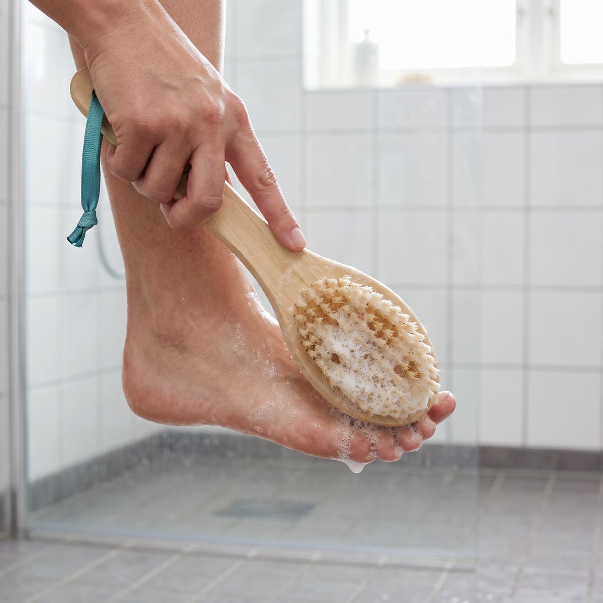 Hand som skrubbar en tvålig fot med en fotborste i trä under duschen i ett kaklat badrum