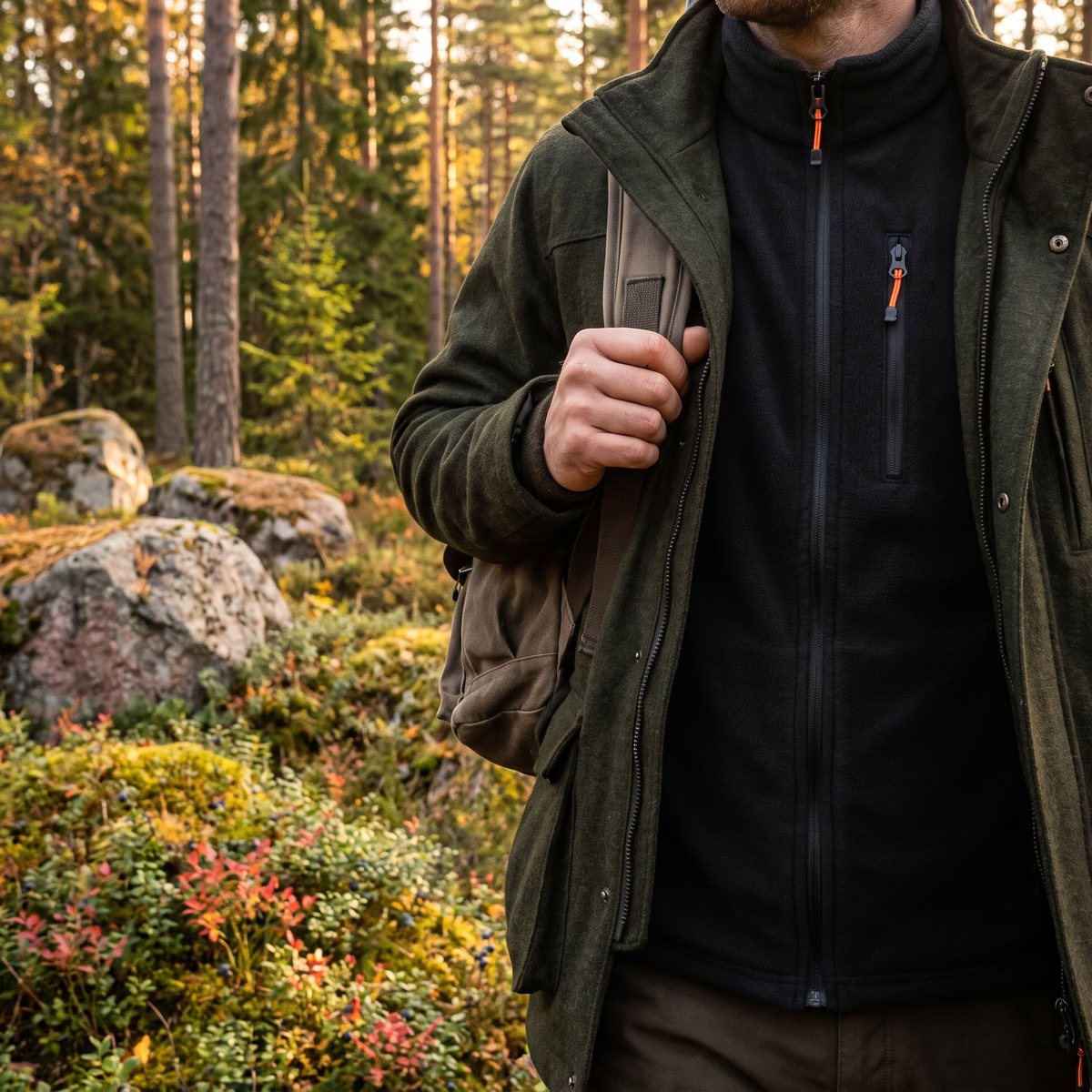 Alpenheat Fire-Fleece, en svart fleeceväst, under en öppen grön jaktjacka på en jägare som går genom svensk tallskog på hösten