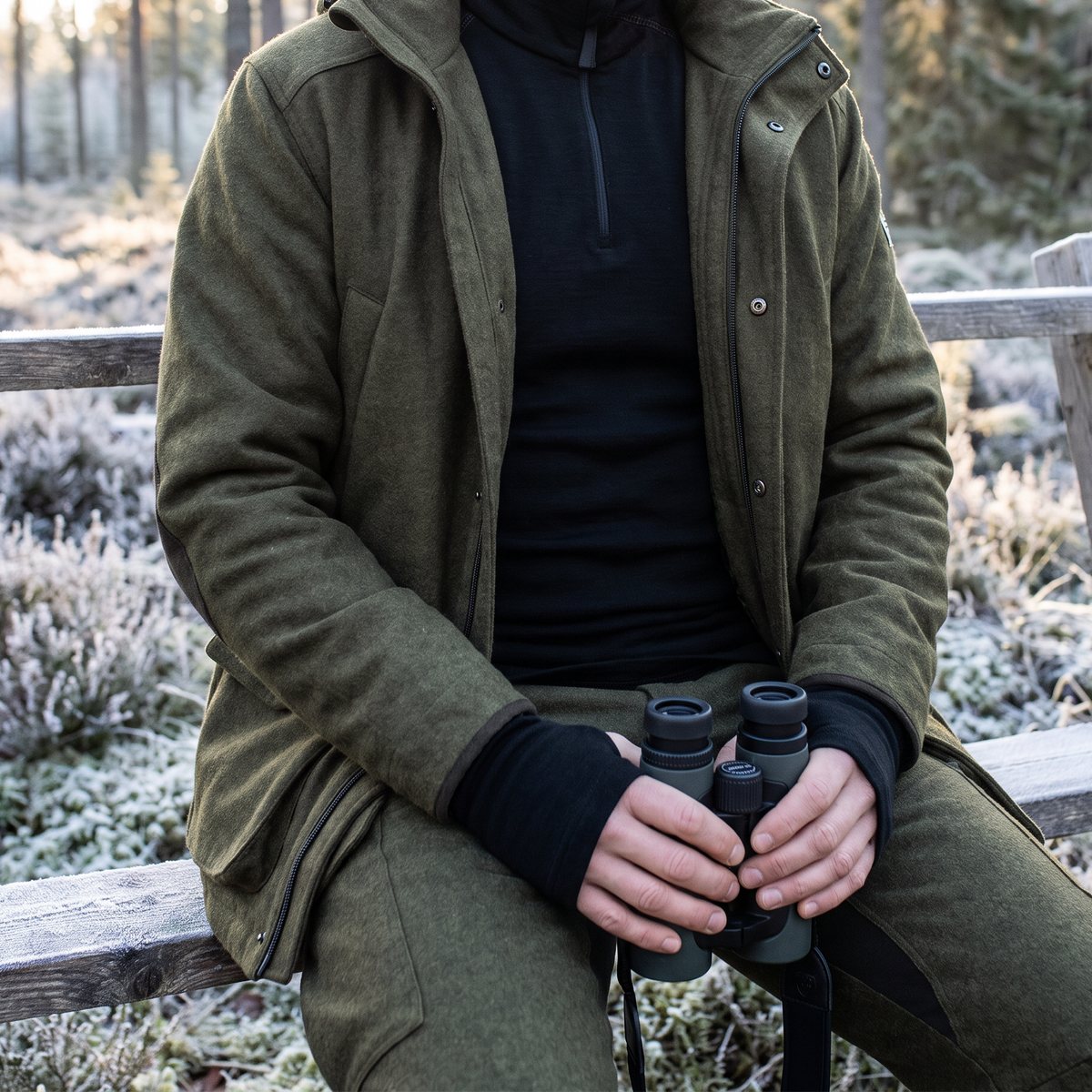 Jägare sitter på en frostig bänk i svensk tallskog med kikare i händerna och svart Devold-tröja med halv dragkedja under öppen jaktjacka