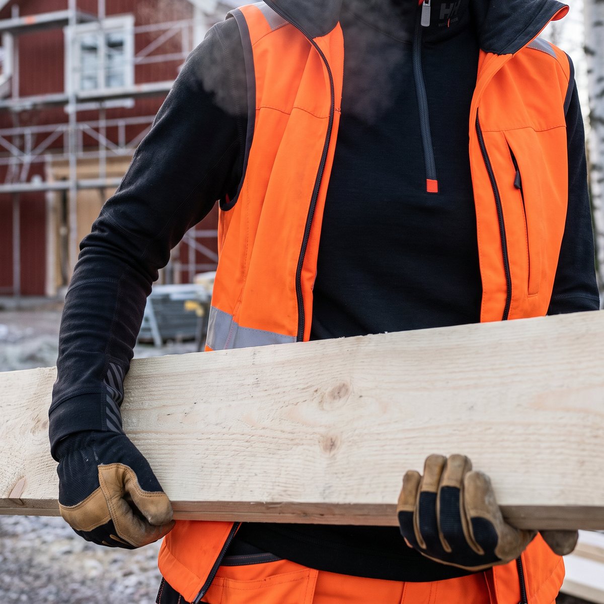 Kvinna i svart Helly Hansen-tröja med halv dragkedja och varselväst bär en planka vid ett rött trähus under bygge