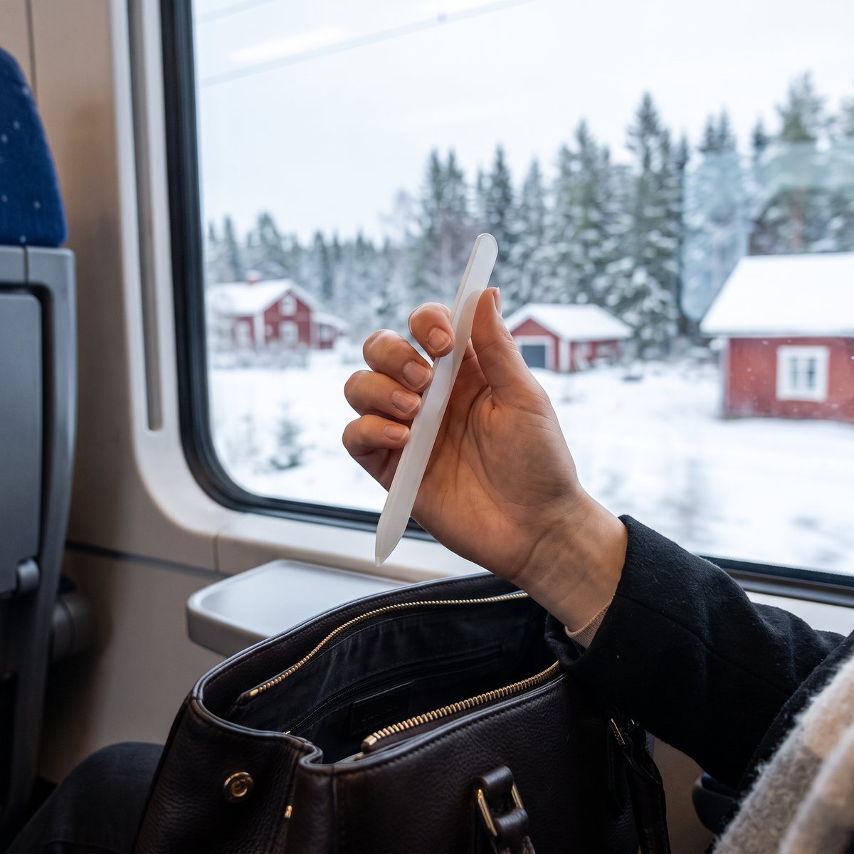 Hand håller en vit glasfil ovanför en öppen handväska vid fönstret på ett tåg genom ett snöigt svenskt landskap med röda stugor