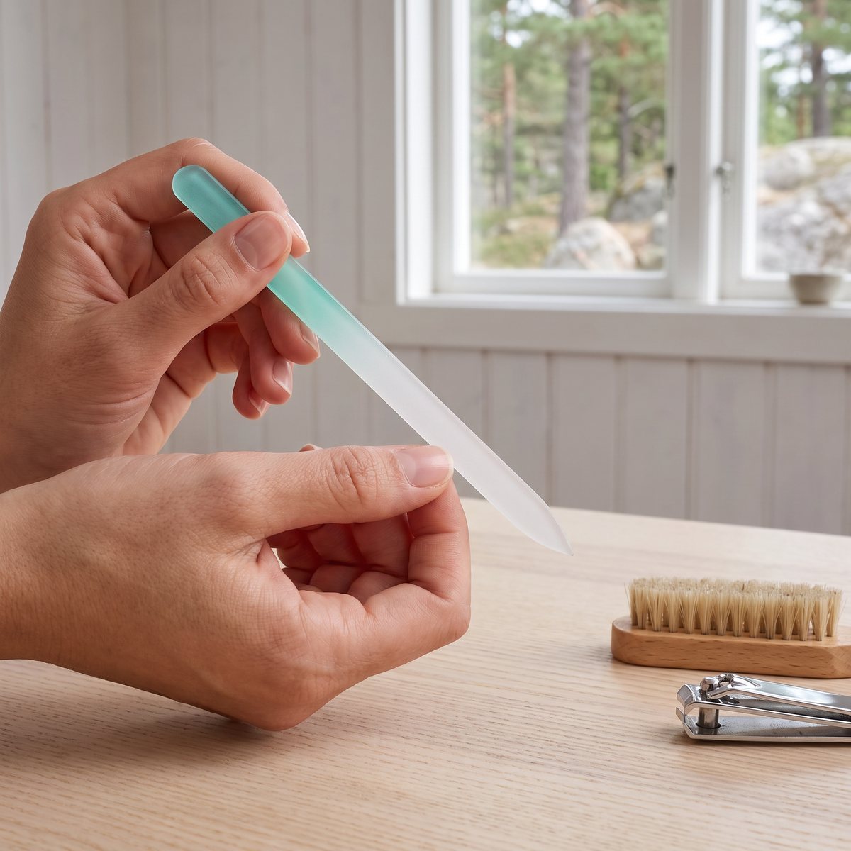 Händer filar en nagel med en mintgrön glasfil vid ett träbord med nagelborste och nagelklippare i en vitpanelad svensk sommarstuga